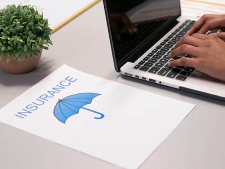 Een persoon typt op een laptop naast een document met een blauw paraplu-symbool en het woord 'INSURANCE', op een bureau met een kleine pot plant.