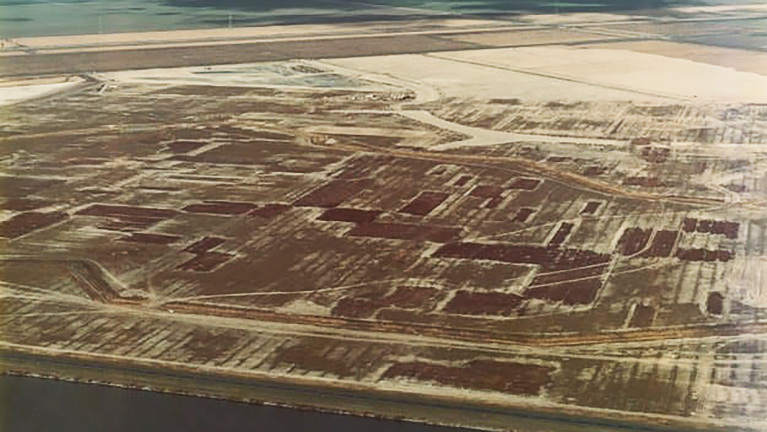 Luchtfoto van een open veld met geometrische vormen, omgeven door wegen en een waterweg aan de onderkant.