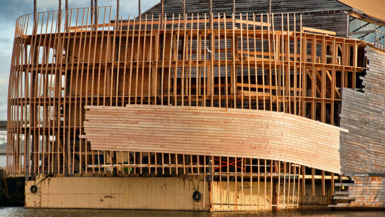 Houten structuur in aanbouw langs een rivier, met verticale en horizontale balken.