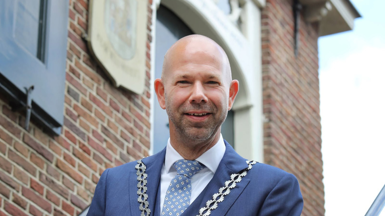 Kale man in blauw pak met ambtsketen, staand voor een bakstenen gebouw
