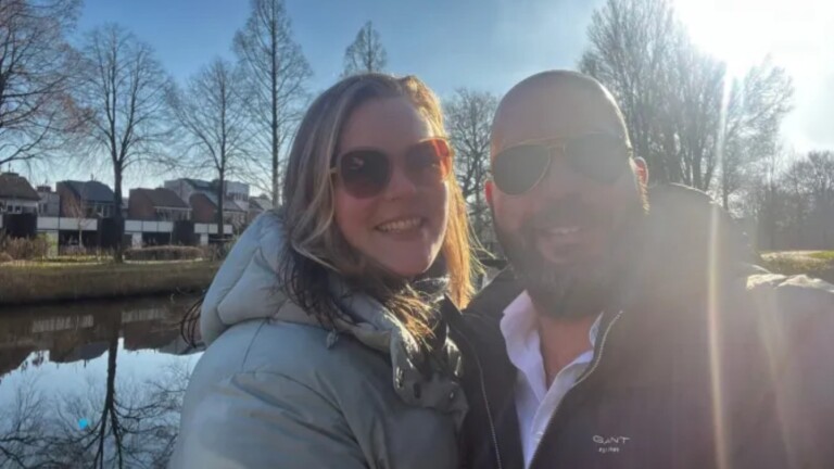 Een lachende man en vrouw met zonnebrillen nemen een selfie in een zonnig park, met bomen en huizen op de achtergrond.