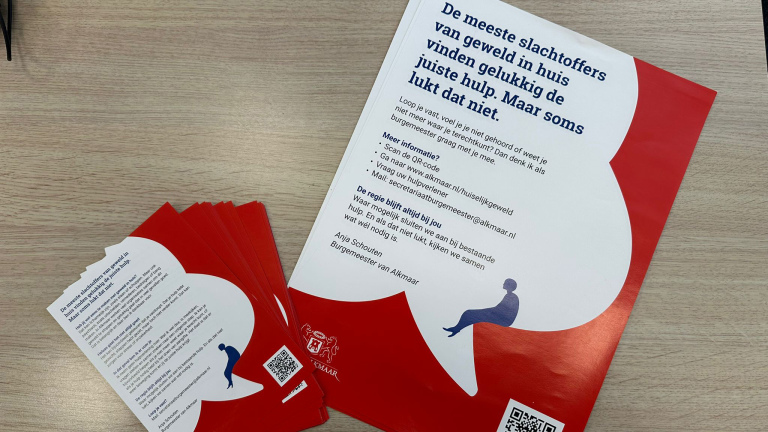 Rode flyers op een tafel met tekst over hulp bij huiselijk geweld in Alkmaar, met contactinformatie en QR-code.