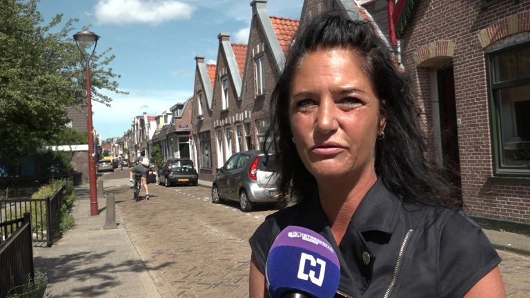 Vrouw in een straat met een rij van bakstenen huizen en een voorbijgangster op een fiets in de achtergrond; de vrouw spreekt in een microfoon met een blauw logo.