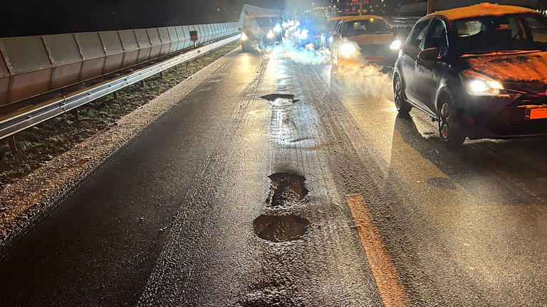 Auto's rijden over een snelweg met meerdere grote gaten in het wegdek, 's nachts verlicht door koplampen.