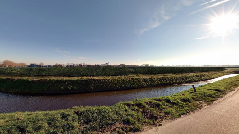 Landschap met een waterkanaal omringd door gras, en in de verte een rij huizen tegen een blauwe hemel met zonnige uitstraling.