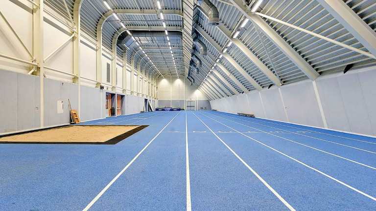 Indoor atletiekbaan met blauwe vloer, hoge plafonds en meerdere banen, naast een zandbak voor verspringen.