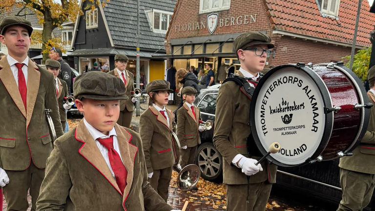 Jonge muzikanten van een trompetterkorps in uniformen met petten en rode stropdassen, staan gereed met hun instrumenten op een straat naast een gebouw met het opschrift "Florian Bergen".