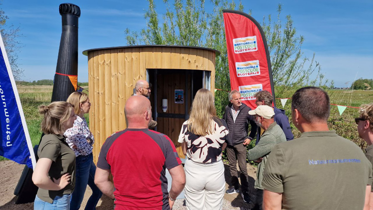 Een groep mensen staat buiten bij een houten gebouwtje, met vlaggen van de Nationale Postcode Loterij.