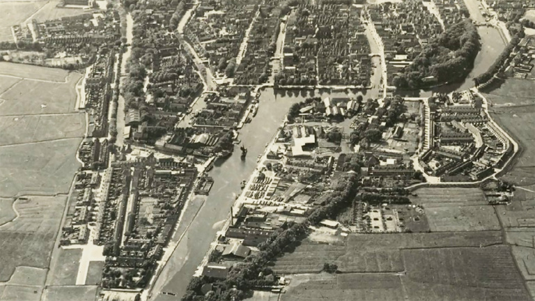 Luchtfoto van een stad met een kanaal en omringende bebouwing en weilanden.