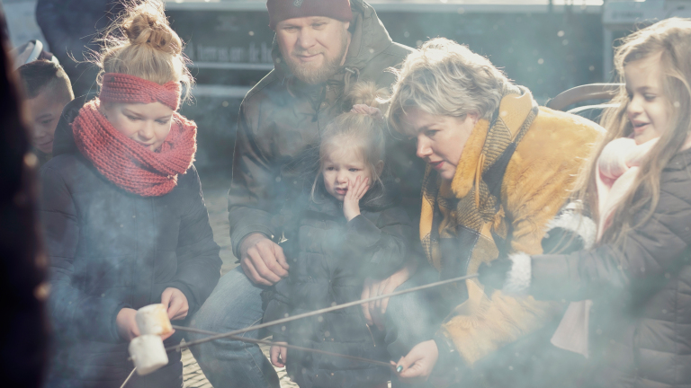 Een groep mensen, waaronder kinderen, roostert marshmallows boven een kampvuur; een warme sfeer met winterkleding en rook zichtbaar.