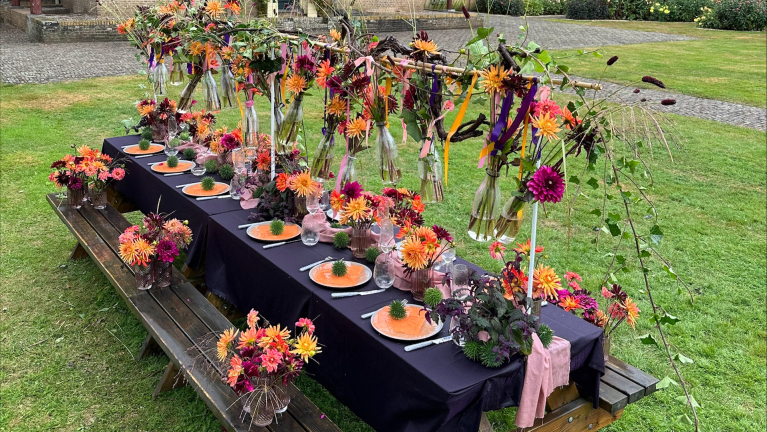 Langwerpige eettafel buiten gedekt met zwart tafelkleed, kleurrijke bloemenarrangementen en borden met oranje decoraties. Boven de tafel hangen vaasjes met bloemen, vastgebonden met linten, en op de tafel staan glazen, servetten en bestek. De omgeving is een grasveld met een bestrate achtergrond.