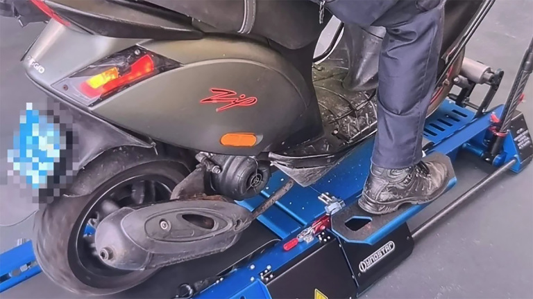 Een persoon staat op een blauwe scooterlift met een Piaggio Zip scooter erop.