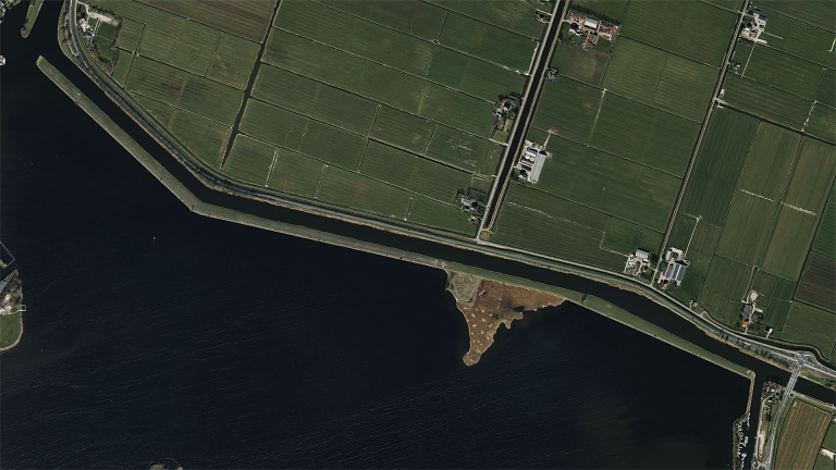 Luchtfoto van een dijk met grasland en water, omgeven door landbouwvelden en enkele gebouwen.