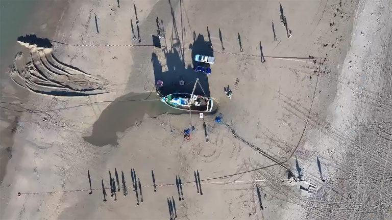 Bovenaanzicht van een strand met een boot, voertuigen en schaduwen van mensen op het zand.