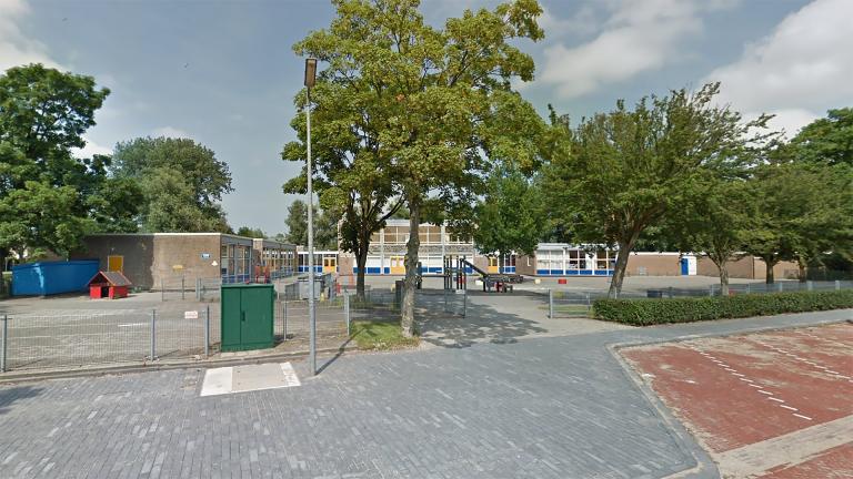 Schoolplein met speeltoestellen en bomen, omgeven door een laag hek.