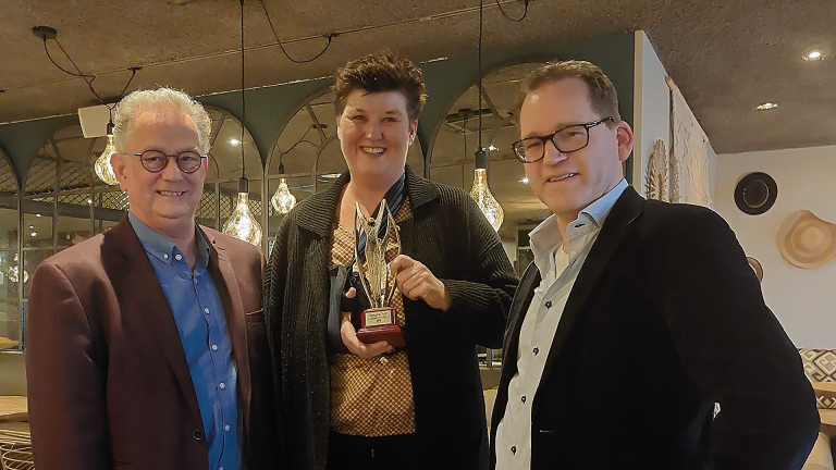 Drie mensen poseren voor een foto in een restaurant, één van hen houdt een trofee vast.