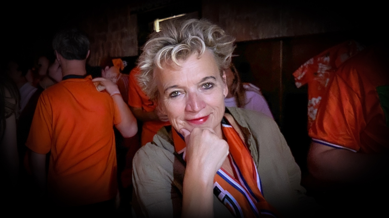 Portret van een blonde vrouw met oranje sjaal, hand onder kin, in een drukke ruimte met mensen in oranje