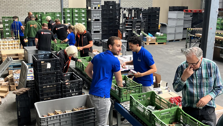 Vrijwilligers werken in een magazijn aan het sorteren van voedsel in groene kratten, met volle schappen en dozen op de achtergrond.