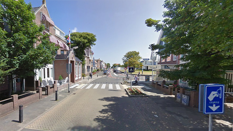 Stadsstraat met zebrapad, geflankeerd door traditionele huizen met rode luifels en groen, met bewegwijzering voor opladen aan rechterkant.