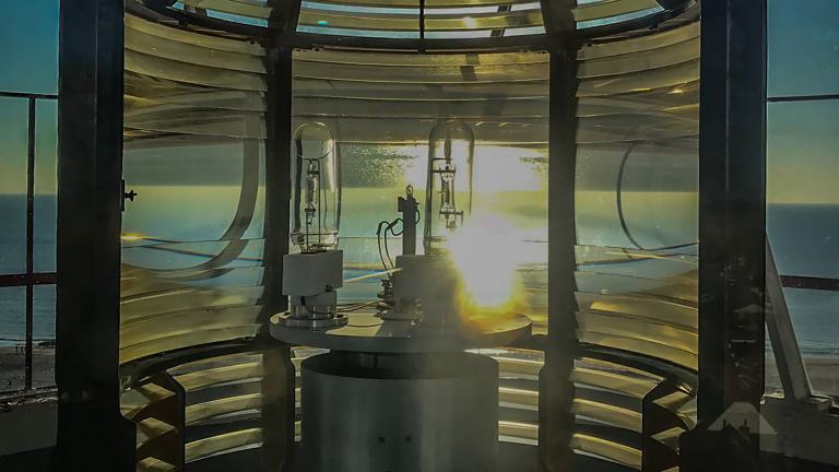 Binnenkant van een vuurtorenlens met zonlicht dat door de glazen panelen valt, met uitzicht op de oceaan.