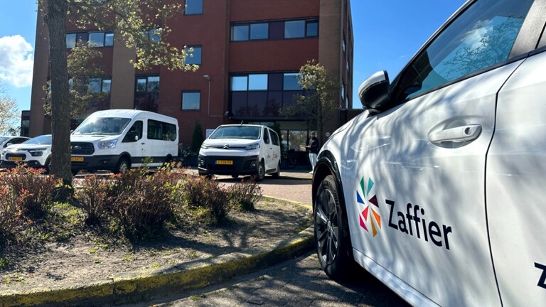 Een witte auto met het logo en de naam 'Zaffier' geparkeerd voor een bakstenen gebouw, met andere voertuigen op de achtergrond onder een blauwe lucht.